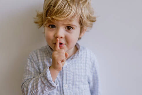 Nettes kleines Kind in Freizeitkleidung steht auf weißem Hintergrund des Studios und legt den Zeigefinger auf die Lippen, während es in die Kamera schaut - ADSF10920