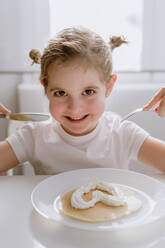 Begeistertes kleines Kind in lässigem T-Shirt sitzt am Tisch mit einem Teller leckerer Pfannkuchen, garniert mit herzförmiger Schlagsahne und schaut fröhlich in die Kamera - ADSF10924