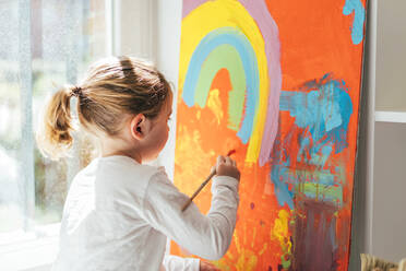 Kreatives blondes Mädchen in Freizeitkleidung sitzt auf der Fensterbank vor dem Fenster und malt mit dem Pinsel einen großen bunten Regenbogen auf orangefarbener Leinwand - ADSF10926