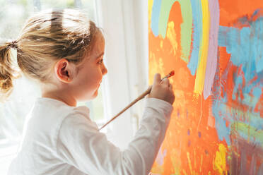 Kreatives blondes Mädchen in Freizeitkleidung sitzt auf der Fensterbank vor dem Fenster und malt mit dem Pinsel einen großen bunten Regenbogen auf orangefarbener Leinwand - ADSF10927