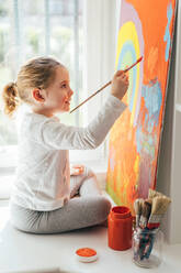 Creative blond girl in casual clothes sitting on window sill against window and painting with paintbrush large multi colored rainbow on orange canvas - ADSF10928