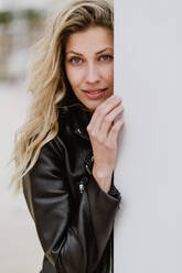 Trendy long haired blonde in black jacket pensively looking at camera standing behind corner of white wall on street - ADSF10932