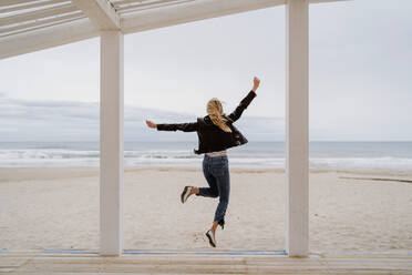 Back view of trendy unrecognizable woman in black jacket merrily jumping with arms raised on white wooden pier with ocean on background - ADSF10933