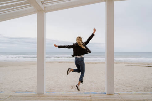 Rückenansicht einer trendigen, nicht erkennbaren Frau in schwarzer Jacke, die fröhlich mit erhobenen Armen auf einem weißen Holzsteg mit Meer im Hintergrund springt - ADSF10933