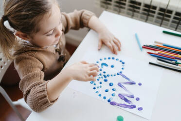 Nettes Kind in Freizeitkleidung malt mit den Fingern, während es am Tisch mit Buntstiften und Scheren in einem hellen Kindergarten sitzt - ADSF10934