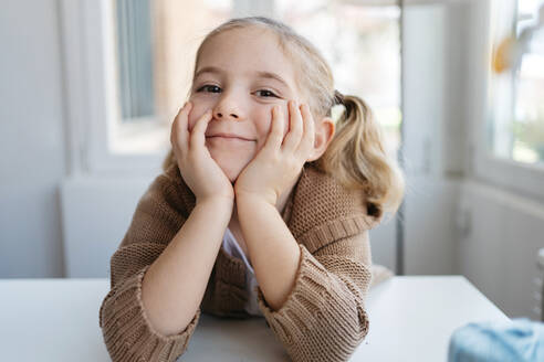 Charmantes kleines Kind in lässigem Pullover, das in die Kamera lächelt und sich auf die Hände stützt, während es am Tisch im hellen Raum des Kindergartens sitzt - ADSF10940