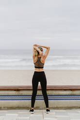 Back view of anonymous female athlete in active wear standing with raised hands in in lock and stretching near seashore on cloudy weather - ADSF10946