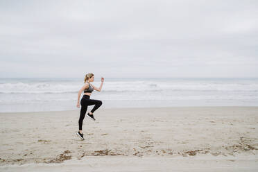 Side view of determined female jogger in stylish sportswear and sneakers doing cardio in jump at empty seashore on cloudy weather - ADSF10949