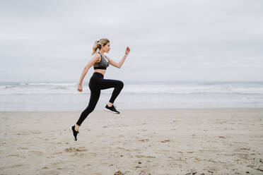 Side view of determined female jogger in stylish sportswear and sneakers doing cardio in jump at empty seashore on cloudy weather - ADSF10950