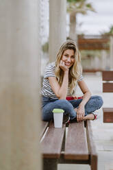 Fröhliche junge Frau, die an einem Sommertag auf einer Bank an der Strandpromenade sitzt und in die Kamera schaut - ADSF10954