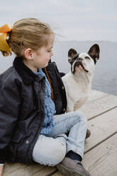 Kleines Mädchen mit französischer Bulldogge sitzt auf Pier in der Nähe von Meer auf trüben bewölkten Tag - ADSF10957