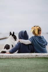 Rückenansicht von zwei Kindern, die eine französische Bulldogge umarmen, während sie gemeinsam am Strand in der Nähe des Meeres sitzen - ADSF10961