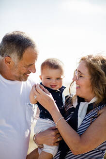 Großeltern lächelnd & halten Enkel am Strand - CAVF88089