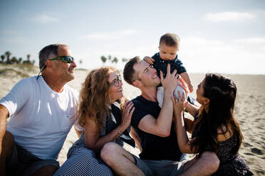Fünfköpfige Familie sitzt am Strand und lächelt ein Kleinkind an - CAVF88092