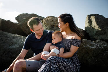 Mitte 30's Eltern halten Sohn auf Felsen am Strand sitzend - CAVF88097