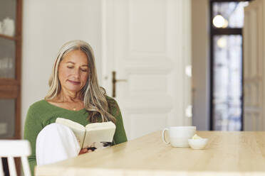 Woman reading book while sitting by table at home - MCF01064