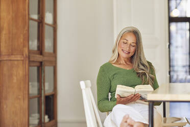Lächelnde Frau liest ein Buch, während sie zu Hause am Tisch sitzt - MCF01069