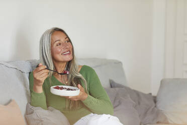 Smiling woman eating breakfast while sitting on sofa at home - MCF01084