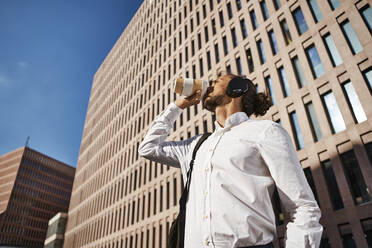 Businessman drinking coffee while listening to music against office building - VEGF02660