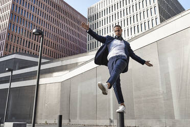 Businessman with headphones balancing on pole against office building - VEGF02661