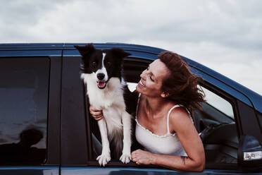 Smiling woman looking at dog while leaning through car window - EBBF00580