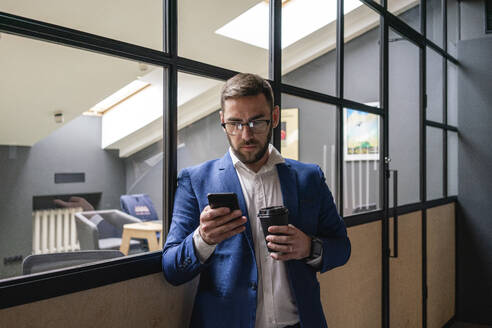 Geschäftsmann hält Einweg-Kaffeebecher, während er sein Smartphone an der Glaswand eines Coworking-Büros benutzt - VPIF02656