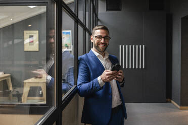 Smiling male professional holding mobile phone and coffee cup while standing by glass wall in office corridor - VPIF02658