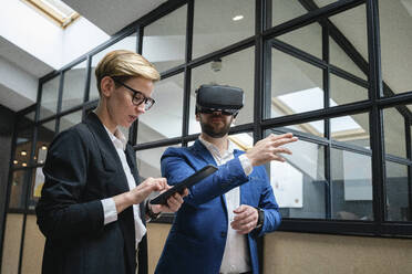 Businesswoman using digital tablet while analyzing colleague experiencing virtual reality simulation at office - VPIF02663