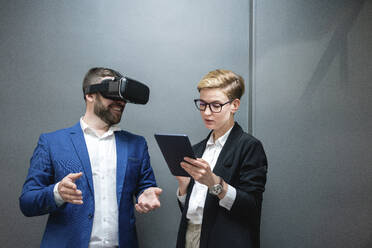Eine Unternehmerin benutzt ein digitales Tablet, während sie einen Kollegen analysiert, der eine Virtual-Reality-Simulation im Büro erlebt - VPIF02664