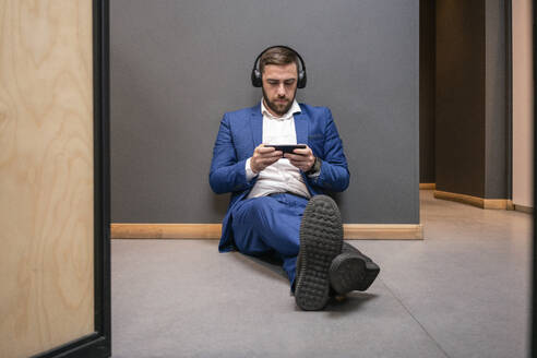 Gutaussehender männlicher Berufstätiger, der ein Videospiel auf seinem Smartphone spielt, während er in der Pause im Korridor des Büros sitzt - VPIF02672