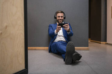 Cheerful male entrepreneur playing video game on smart phone while sitting in corridor at coworking space during break - VPIF02673