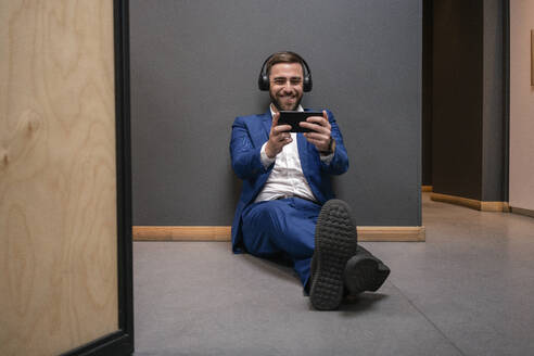 Fröhlicher männlicher Unternehmer, der ein Videospiel auf seinem Smartphone spielt, während er während der Pause im Flur eines Coworking Space sitzt - VPIF02673