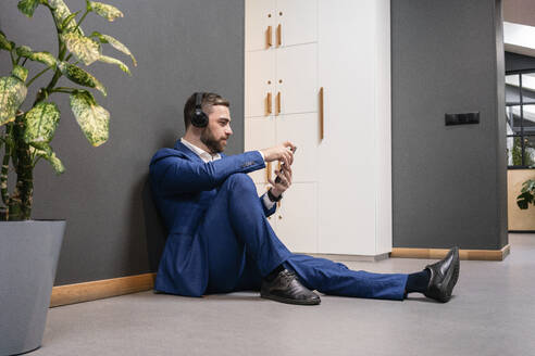 Gutaussehender männlicher Unternehmer, der eine Pause macht, während er Musik über Kopfhörer hört und sein Smartphone in einem kreativen Bürokorridor benutzt - VPIF02675
