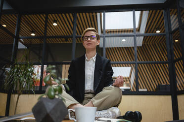 Female professional meditating while practicing yoga on table at coworking office space - VPIF02680