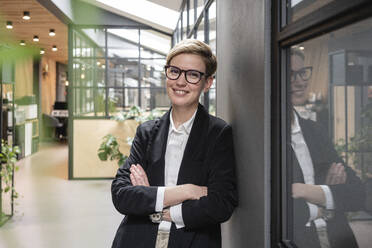 Beautiful female entrepreneur with arms crossed leaning on wall at coworking office corridor - VPIF02689