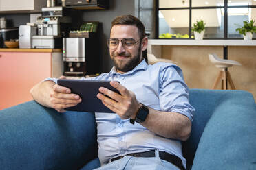 Lächelnder Geschäftsmann, der ein digitales Tablet für einen Videoanruf benutzt, während er in der Büro-Cafeteria auf einem Sessel sitzt - VPIF02718