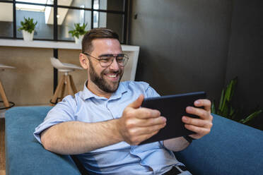 Glücklicher gutaussehender männlicher Fachmann, der ein digitales Tablet für einen Videoanruf benutzt, während er auf einem Sessel in einer Büro-Cafeteria sitzt - VPIF02719
