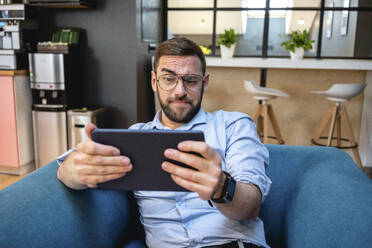 Geschäftsmann mit hochgezogener Augenbraue, der ein digitales Tablet für einen Videoanruf benutzt, während er auf einem Sessel in einer Büro-Cafeteria sitzt - VPIF02722