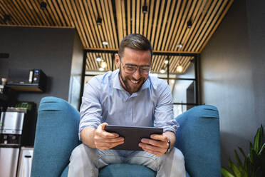 Fröhlicher männlicher Berufstätiger, der ein digitales Tablet für einen Videoanruf benutzt, während er auf einem Sessel in einer Büro-Cafeteria sitzt - VPIF02725