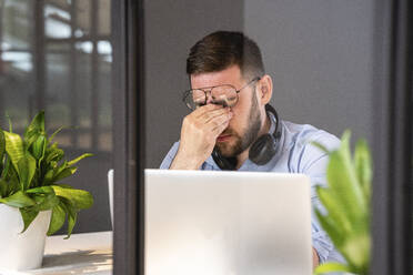 Müde gutaussehende männliche Fachkraft mit Laptop durch Glaswand in kreativem Coworking-Büro gesehen - VPIF02749