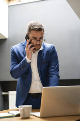 Geschäftsmann, der mit einem Smartphone spricht, während er einen Laptop am Schreibtisch in einem kreativen Büro benutzt - VPIF02750