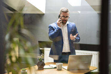 Male entrepreneur talking on smart phone while looking laptop on desk in creative office - VPIF02752