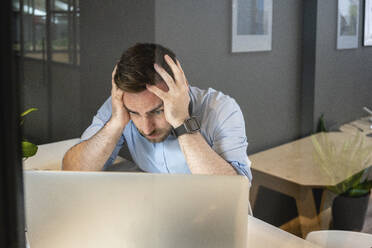 Tired businessman with head in hands looking at laptop on desk in creative coworking office - VPIF02756