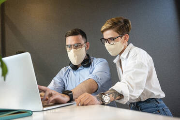 Ein männlicher und ein weiblicher Kollege planen eine Strategie, während sie über einen Laptop am Schreibtisch im Büro während einer Pandemie diskutieren - VPIF02760