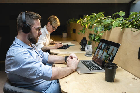 Geschäftsmann bei einem Videogespräch mit Kollegen über einen Laptop, während er neben einer Geschäftsfrau am Schreibtisch in einem Kreativbüro sitzt - VPIF02762