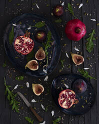 Draufsicht auf frisch geschnittene Feigen und Granatäpfel, die auf Tellern neben Rucola und Messer auf dem Holztisch liegen - ADSF10971