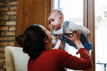 Entzückte Frau, die ein süßes Baby hochhebt und spielt, während sie auf einem Stuhl in der Nähe des Fensters in einem gemütlichen Zimmer zu Hause sitzt - ADSF10974