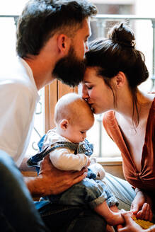Glückliche Eltern küssen Baby und einander beim Sitzen in der Nähe von Fenster in gemütlichen Raum in Wochenende zu Hause - ADSF10977