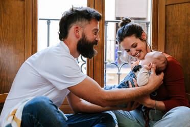 Bearded father and happy mother communicating with cute baby while sitting on floor near window at home - ADSF10978