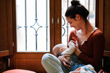 Fröhliche Frau umarmt und stillt süßes Baby, während sie am Fenster sitzt und sich in einem gemütlichen Zimmer zu Hause ausruht - ADSF10979